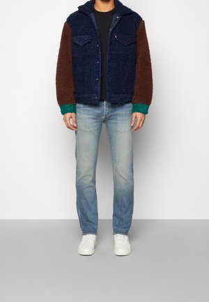 Man draagt een marineblauw en bruin fleecejack met groene manchetten, een zwart shirt, lichtblauwe spijkerbroek en witte sneakers, staand tegen een witte muur.