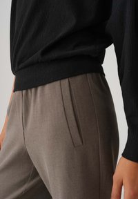 Schwarzes, strukturiertes Oberteil kombiniert mit taupefarbenen, plissierten Hosen, die eine glatte Stoffoberfläche und eine maßgeschneiderte Passform hervorheben. Minimalistische Designdetails sind sichtbar.