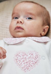 Valkoinen vauva-asuste, jossa on vaaleanpunainen brodeerattu sydämen muotoilu ja aaltoileva kaulus. Ominaisuuksiin kuuluu pehmeä kangas ja nappikiinnitys.