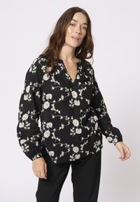 Blusa nera con stampa floreale, scollatura a V, maniche lunghe a sbuffo e polsini elasticizzati. Presenta motivi floreali bianchi su tutta la superficie.