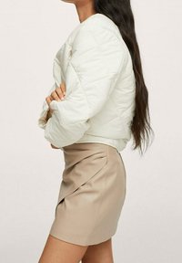 Giacca bomber trapuntata color crema con colletto rotondo, abbinata a una gonna aderente color tan con un design drappeggiato e dettagli a piega laterale.