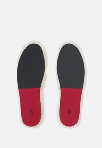 Svarte såler med en rød hælseksjon, med en myk overflate og preget "UGG"-logo på hælen.