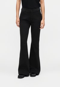 Γυναίκα με μαύρο παντελόνι καμπάνα, μαύρο τοπ και μαύρα γυαλιστερά μυτερά παπούτσια, που στέκεται μπροστά σε ένα απλό φωτεινό φόντο.