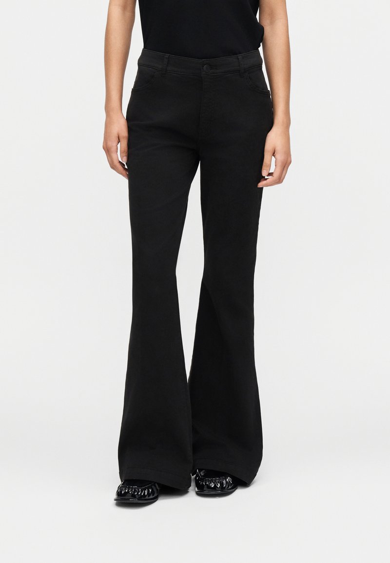 Γυναίκα με μαύρο παντελόνι καμπάνα, μαύρο τοπ και μαύρα γυαλιστερά μυτερά παπούτσια, που στέκεται μπροστά σε ένα απλό φωτεινό φόντο.