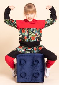 Kolorowy dres z tropikalnym kwiatowym wzorem w zielonym, czerwonym i czarnym kolorze. Dziecko prezentujące mięśnie, siedząc na miękkim niebieskim bloku.