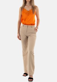 Kobieta stoi w pomarańczowej bluzce bez rękawów z koronkowymi wykończeniami, beżowych spodniach z wysokim stanem, czarnych sandałach na obcasie z odkrytymi palcami oraz minimalną biżuterią.