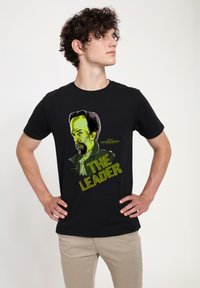 Černé bavlněné tričko s grafickým potiskem obličeje postavy v zelené a žluté barvě, s nápisem "VEDOUCÍ" tučnými písmeny.