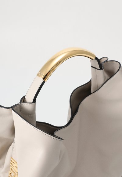 Beige leather tote bag with a ruffled design and a gold-tone arch handle. Features black trim and a textured finish.