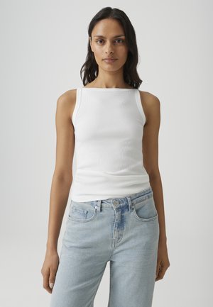 Donna con capelli lunghi e scuri indossa un top bianco senza maniche a coste e jeans azzurri a vita alta, in piedi davanti a uno sfondo chiaro e uniforme.