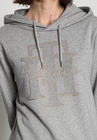 Grijze hoodie van zacht materiaal, met een groot, versierd logo met strass-steentjes en gouden accenten aan de voorkant. Met trekkoord in de capuchon.