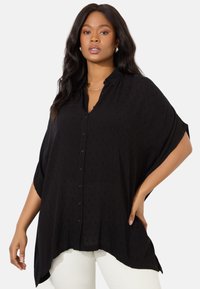 Svart skjorta med knappar, avslappnad passform, korta ärmar, texturerat tyg och rundad nederkant, i kombination med vita byxor.