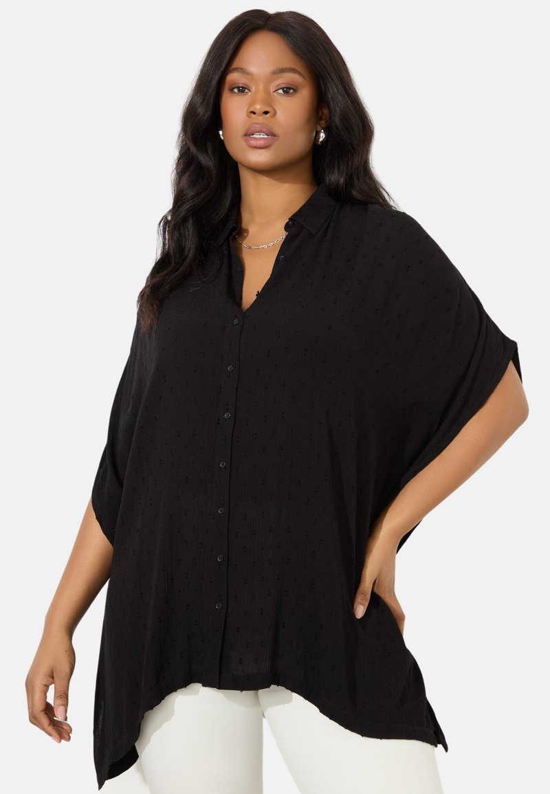 Svart skjorta med knappar, avslappnad passform, korta ärmar, texturerat tyg och rundad nederkant, i kombination med vita byxor.