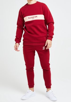 Sweatshirt rouge avec une bande horizontale crème et le texte "ILLUSIVE LONDON"; joggeurs rouges assortis avec broderie; associés à des baskets blanches.