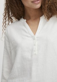 Camicia bianca in lino con scollo a V e cinque bottoni. Presenta maniche lunghe e un tessuto leggermente texturizzato. Design minimalista.