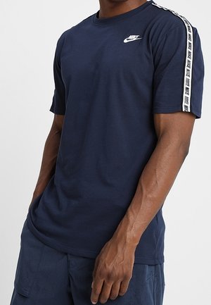 Mann trägt ein dunkelblaues Nike-T-Shirt mit weißem Logo auf der Brust und einem Markenstreifen entlang der Schulter und des Ärmels, kombiniert mit dunkelblauen Shorts.