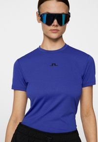 Royalblå, figurnära träningströja med korta ärmar och rund halsringning, prydd med en liten svart logotyp på bröstet. Strukturerad, elastisk tyg.