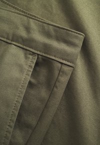 Olivgrüner Stoff mit einer glatten, strukturierten Oberfläche. Verfügt über eine auffällige Tasche und kontrastierende Nähte. Robustes und strukturiertes Design.