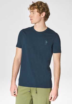 Giovane uomo con capelli ricci che indossa una maglietta blu navy a girocollo e pantaloni verde oliva, guarda a sinistra contro uno sfondo chiaro e uniforme.