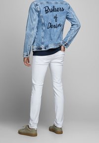 Osoba stojí s jednou rukou v kapse, na sobě má světle modrou denimovou bundu s textem "Brothers of Denim", bílé kalhoty a béžové boty.