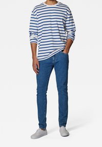 Langärmliges weißes Hemd mit blauen horizontalen Streifen, kombiniert mit blauen Jeans. Modell trägt weiße Sneakers, Hände in den Taschen.