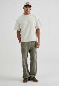 Tricou oversized bej deschis, cu mâneci scurte; pantaloni de trening verzi măslinii cu buzunare laterale; încălțăminte maro. Design minimalistic, fără modele.