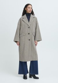 Donna che indossa un lungo cappotto a scacchi beige e blu marino sopra un dolcevita nero, jeans a gamba larga e stivali neri con tacco, in piedi contro uno sfondo neutro.