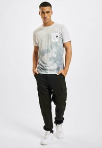 Valge T-särk, millel on peen sinine tie-dye muster, koos rinna taskuga. Kombineeritud mustade cargo-pükstega ja valgete tennistega.