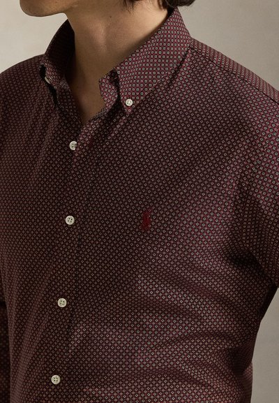 Camisa abotoada bordeaux com um padrão geométrico, apresentando um colarinho clássico e pequenos botões brancos. Logótipo vermelho no peito esquerdo.