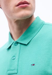 Κοντινό πλάνο ενός ατόμου που φοράει γαλάζιο πόλο μπλουζάκι με κουμπιά και  ένα μικρό κεντημένο λογότυπο σε κόκκινο, λευκό και μπλε στο στήθος.