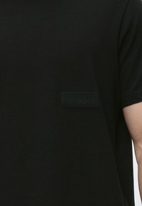 Chemise noire à manches courtes avec un petit patch rectangulaire en caoutchouc sur la poitrine affichant "BOSS" en lettres noires assorties.