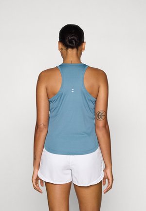 Frau mit Haarknoten, die ein blaues Racerback-Top und weiße Sportshorts trägt, steht mit dem Rücken zum Betrachter vor neutralem Hintergrund.