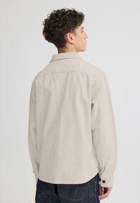Chemise beige clair en tissu doux, dotée d'un col italien et de boutons aux poignets. Le dos présente un design simple sans logo.