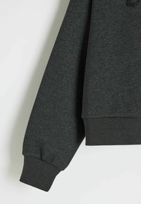 Ciemnozielony sweter z teksturowanym wykończeniem, z ribbowanymi mankietami i dołem oraz luźnym krojem. Tkanina ma miękki wygląd.