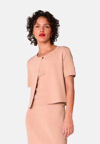 Hellrosa, figurbetontes Oberteil mit kurzen Ärmeln, rundem Ausschnitt und einem zentralen goldfarbenen Verschlussdetail. Hergestellt aus einem weichen, strukturierten Stoff.