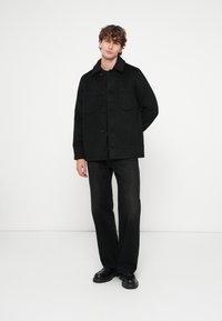 Sort skjortejakke med to brystlommer, lavet af tekstureret stof. Matcher med mørke jeans og sort fodtøj, stående mod en ensfarvet baggrund.