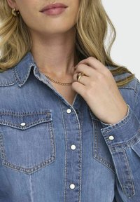 Blonde Frau, die ein blaues Jeanshemd mit weißen Knöpfen, eine goldene Kettenhalskette, einen Ring und kreisförmige Ohrringe trägt und ihre Halskette anpasst.