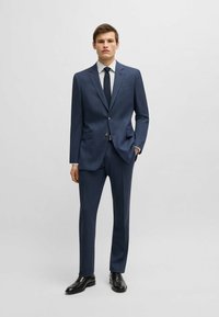 Navyblauwe pak met een gestructureerd patroon, gecombineerd met een wit overhemd met kleine zwarte stippen en een bijpassende stropdas. Zwarte nette schoenen.