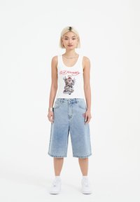 Valkoinen hihaton paita, jossa punainen graafinen kuvio, yhdistettynä vaaleansiniin farkku-Bermuda-shortsit. Molemmissa vaatteissa on rento istuvuus ja epämuodollinen tyyli.