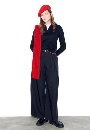 Kvinde iført sort langærmet top, brede sorte bukser, sorte sko, rød beret og rød tørklæde draperet over skulderen, stående på betongulv.