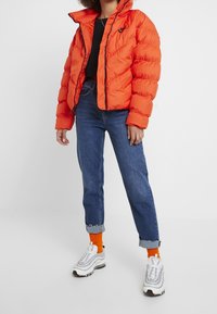 Orange puffer jacket with a zip front and black logo, worn over a black top, paired with light blue rolled jeans and white sneakers.