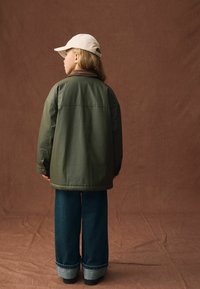 Zielona oliwkowa kurtka ocieplana z brązowym kołnierzem, noszona na ciemnoniebieskich spodniach dżinsowych o szerokich nogawkach z podwiniętymi dolnymi końcami. Jasny czapka baseballowa.