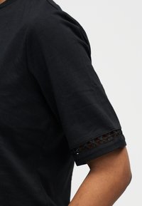 Gros plan d'une personne portant une chemise noire à manches courtes avec une bordure en dentelle florale près du poignet.