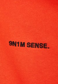 Oranžinė megzta medžiaga su juodu siuvinėjimu "9N1M SENSE." su glotnia tekstūra ir vienodu raštu.