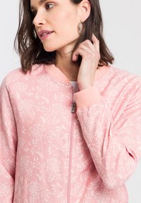 Jasnoróżowa bomberka z całkowitym białym wzorem kwiatowym, prążkowanymi mankietami, zamek błyskawiczny oraz luźnym krojem.