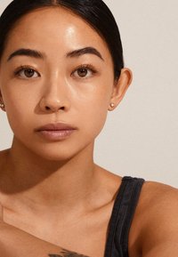 Persona con tono di pelle medio e capelli neri lisci, che indossa un top senza maniche. Piccole orecchini d'oro. Pelle liscia con trucco naturale.