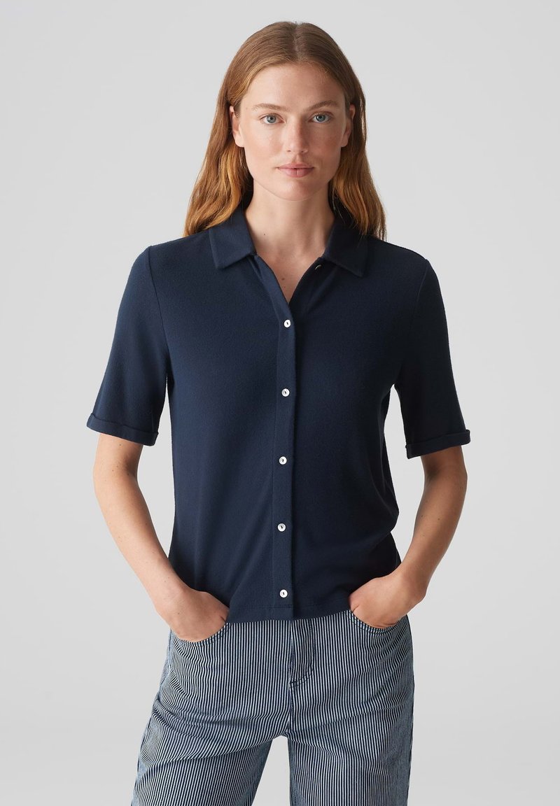Camicetta blu navy a maniche corte con bottoni, realizzata in un tessuto liscio, con colletto e polsini arrotolati, abbinata a pantaloni a righe.