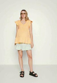 Top laranja de mangas curtas com decote em V e bainha em camadas, combinado com uma saia mini verde-clara com babados. Sandálias pretas completam o look.