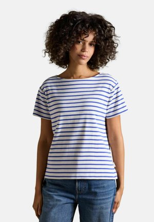 Kobieta o kręconych włosach, ubrana w biały krótki rękaw z niebieskimi poziomymi paskami i niebieskie dżinsy, stojąca na jednolitym tle.