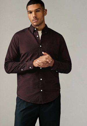 Camisa de botones burdeos con mangas largas y un bolsillo en el pecho, que presenta botones blancos y una tela texturizada. Lleva puesta sobre una camisa clara.