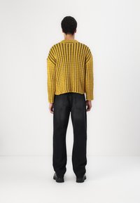 Żółty sweter na drutach w czarne paski, o luźnym kroju i prążkowanej fakturze. Noszony z czarnymi szerokimi spodniami dżinsowymi i czarnymi masywnymi butami.